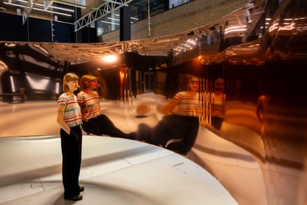 A person standing looking at an installation of a shiny, bending piece of metal spanning the room