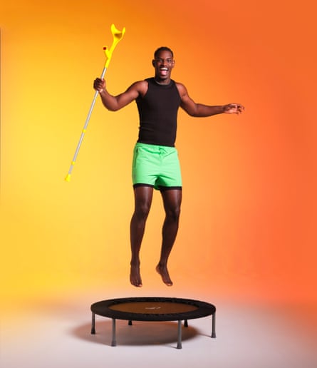 A man wearing green shorts and a black vest, jumping on a small trampoline while holding a crutch, shot against a graduated yellow and orange background.
