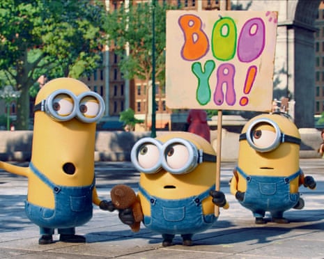 Three Minions in denim overalls stand on a street holding a colorful 'BOO YA!' sign