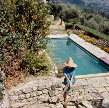 A woman in a straw hat, blue top and capri pants sits on a stone wall with her back to a swimming pool that is surrounded by bushes and flowerbeds