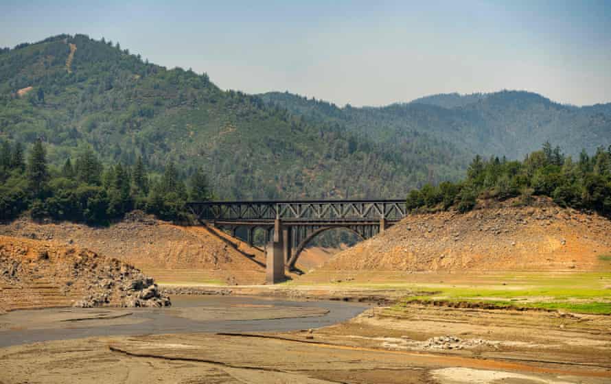 Forested mountains overlook a dry lakebed. A trestle bridge over mud and rock connects two hills.