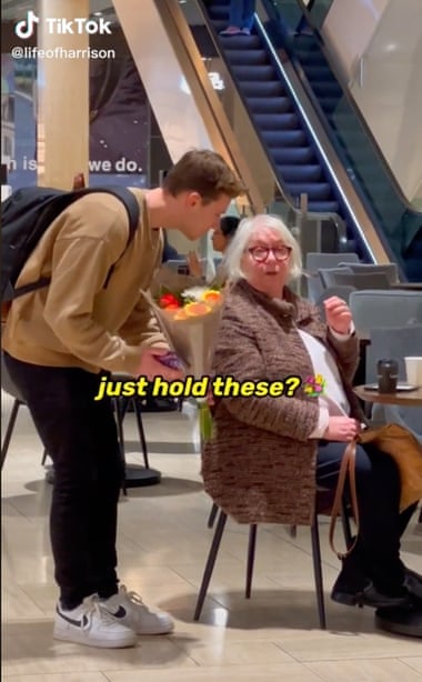 A screenshot of Harrison Pawluk's TikTok video showing him leaning over Maree, who is sitting in a cafe, asking her to hold a bouquet of flowers.