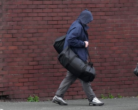 A man walks along a path next to a high brick wall carrying a duffel bag