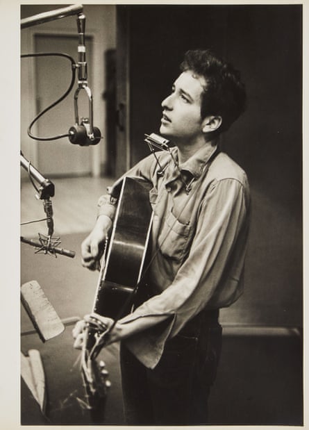 Bob Dylan, in a long-sleeved shirt with a collar and something in a front pocket, strums a guitar and sings into a hanging microphone in a 1961 photograph
