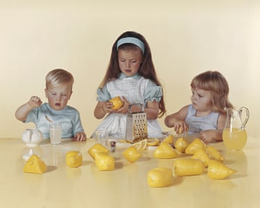 Colour photograph of three children sat at a table with lemons spread out, along with a grater and a jug
