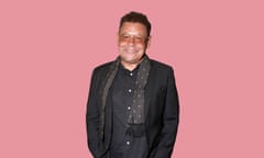 Craig Charles wearing a black suit and tie, glasses and a black scarf with white spots, against a pink background