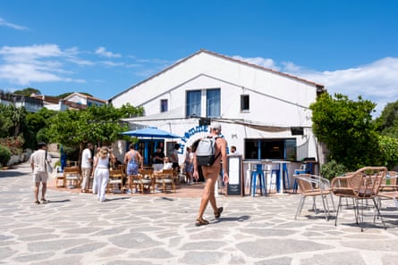 A restaurant in a pedestrian square, with some clothed people outside and one man wearing just a hat, backpack and shoes