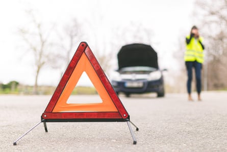 A woman talking on phone while her car is broken down on the road with a reflective warning triangle in front of it.