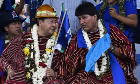 Two middle-aged Bolivian men, in matching ceremonial robes of dark red with orange lines and adorned with white garlands of flowers, appear to chat with one another outdoors seated in chairs.