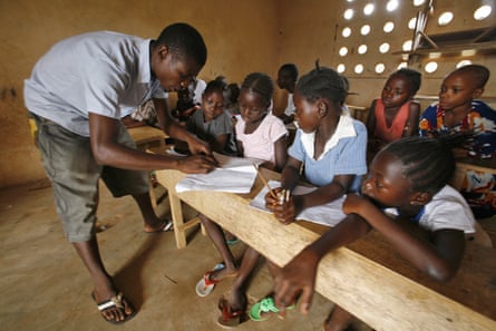 A teacher leans in to look at pieces of paper on a wooden table while students look on