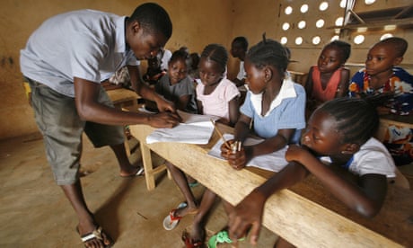 A male teacher with a class of children sitting at wooden desks in Sierra Leone