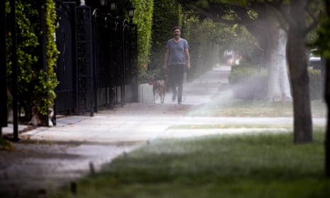A man walks a dog down a sidewalk as sprinklers water patches of grass and trees next to the sidewalk.