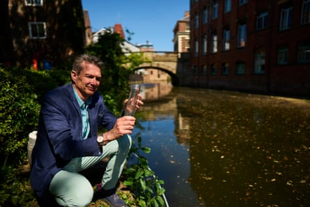 A man crouches next to a river with a bridge and buildings in the background.