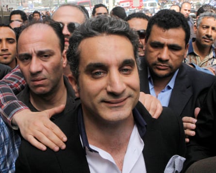 A closeup of Bassem Youssef shielded by two men behind his as they move through a crowd of people