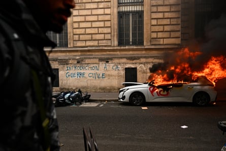 A car burns in a Paris street near graffiti on a stone wall reading ‘introduction to civil war’. Flames and black smoke are pouring from its windows. A soldier stands in shadow in the foreground.