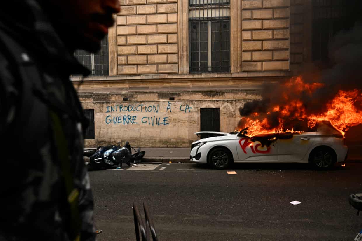 A car burns in a Paris street near graffiti on a stone wall reading ‘introduction to civil war’. Flames and black smoke are pouring from its windows. A soldier stands in shadow in the foreground.