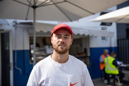 A man in a white T-shirt and red cap, standing in front of a takeaway sandwich shop