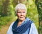 Dame Judi Dench, with short white hair and a blue scarf, stands outdoors among green trees