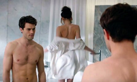 Jamie Dornan, as Christian Grey in Fifty Shades of Grey, is pictured bare-chested in front of a mirror, with a partially dressed woman in the background
