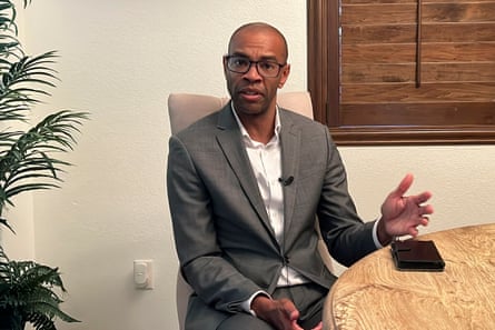 A man wearing a suit sits at a table while speaking.