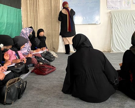 Eight girls in headscarves sit on the floor and take notes while another stands at a whiteboard writing