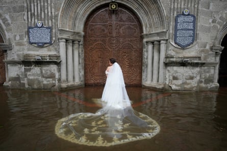A woman in a wedding dress stands in water outside the door of a church, her long veil floating.