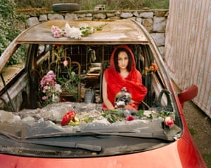 A portrait of Farah, Aabey, Lebanon, 2020, shows Farah wearing red and sitting in her burned out car, which is strewn with flowers