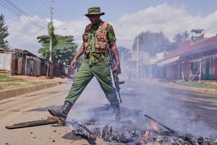 An anti-riot police officer wearing khaki trousers and hat and big black boots kicks burning debris. He carries a gun.