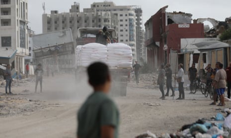 People watch a truck driving past damaged buildings carrying large white sacks of supplies