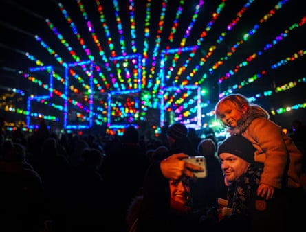 A woman takes a selfie of a child on a mans shoulders with 2026 light show in background