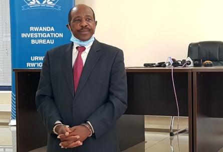 Paul Rusesabagina in a suit and tie, handcuffed, wearing a face mask pulled down around his chin, in front of a sign saying ‘Rwanda Investigation Bureau’