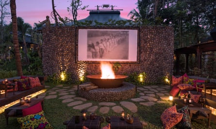 A screen mounted on a wall with the sky behind, a fire pit in front and low-level seating with bright cushions around