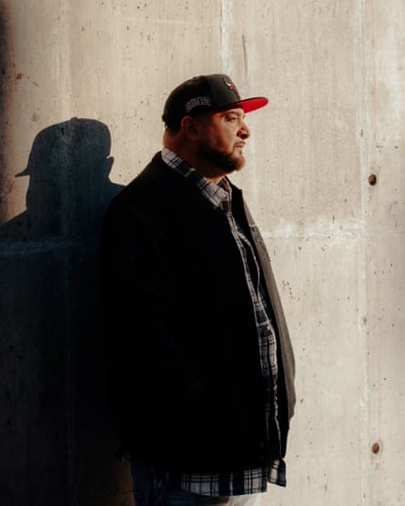 Edgardo Colon pictured from the thighs up, side-on to the camera, looking to his left, wearing a checked shirt, a dark jacket and a baseball cap, his silhouette on the wall he’s leaning against