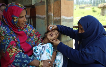 A woman holds a child while another woman administers a drop of vaccine into their mouth