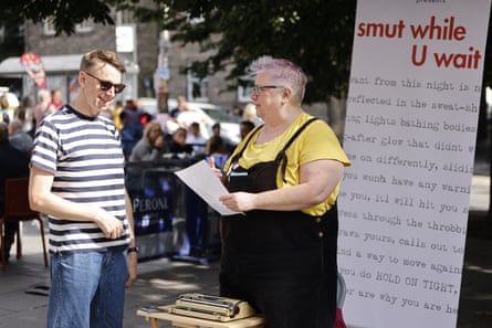 A woman in bib and yellow t-shirt holding a piece of paper in front of a billboard stating, ‘Smut While U Wait’. A man in a stripy T-shirt and glasses is talking to her