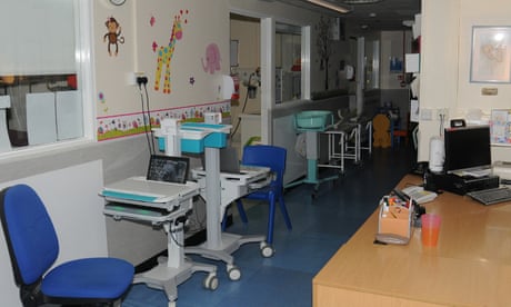 A hospital corridor with a desk and mobile tables with computers and chairs, with rooms coming off the side