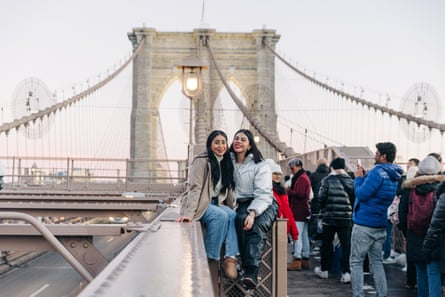A crowd of people walk on a path over the Brooklyn Bridge while two young women pose for a photo.