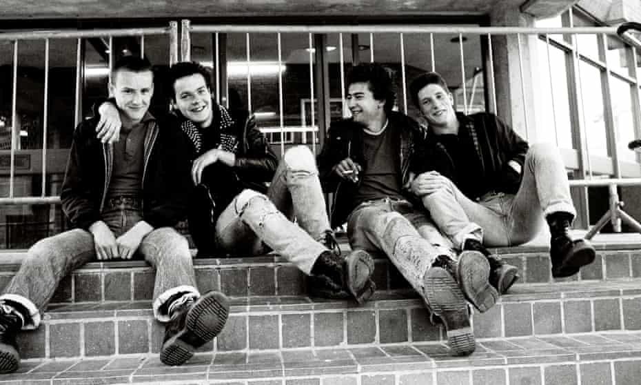 Black and white image of four young white men in leather jackets and ripped jeans sitting on steps smiling one with arm round the other.