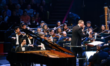 Pianist Alim Beisembayev at a grand piano, behind conductor John Wilson who is surrounded by the string players of the Sinfonia of London on stage at the Royal Albert Hall