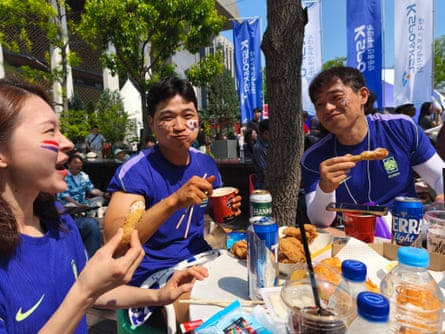 Three people in blue shirts sitting and eating with food and drink spread round a table