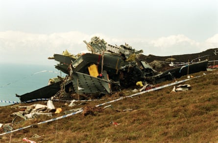 The wreckage of the crashed helicopter strewn across a clifftop hillside, with the sea in the background. The accident scene is cordoned off by police tape.