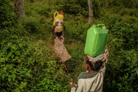 Children with containers balanced on their heads make their way along a path lined with thick undergrowth.