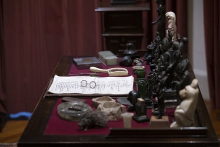 a tabletop or desk surface with a collection of black and white figurines which are arranged in lines like chess pieces, other artefacts and ornaments, a magnifying glass and a sheet of paper with Freud’s spectacles placed on it.