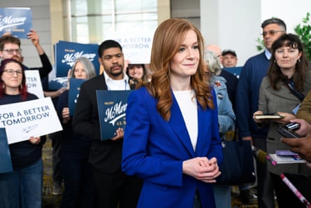 A woman with long hair in a bright blue blazer, backed by supporters with signs, smiles at tape recorders being held in front of her.