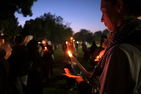 A man holds a lit candle in cupped hands at an outdoor evening vigil with others in the background