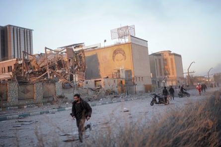 A man runs towards the left of the picture, past a destroyed building behind. A mural of the ayatollah is on a wall of an adjacent building
