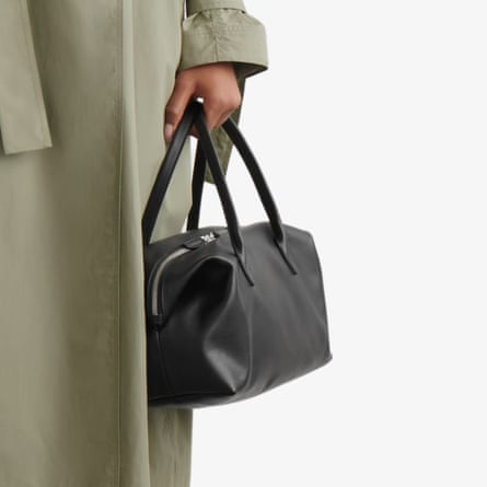 A model holds the Montmarte bowling bag in black. The model is wearing a sage raincoat the image is a close up of the hand holding the bag.