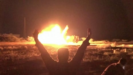A man with his arms in the air as he watches an explosion in the distance.