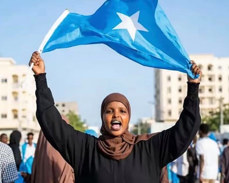A young woman in a hijab stands holding a Somali flag above her head, shouting