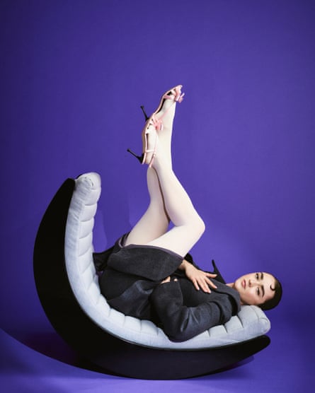 Maisie Williams looking at the camera and lying on a curved grey and black sofa seat with her legs up in the air, touching at ankles, wearing a dark blazer and skirt, white tights and pale-pink stilettos, against a deep-purple background